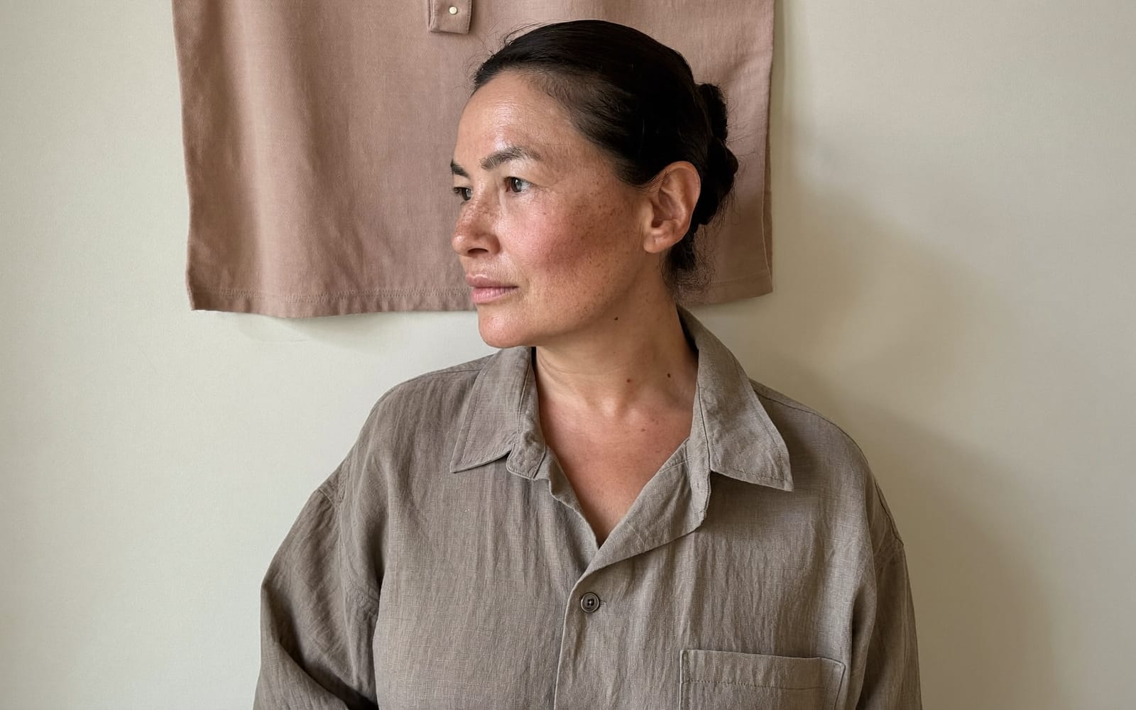 A person in three-quarter profile, looking away from the camera, in soft daylight against an ivory wall.